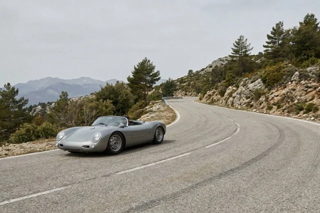 A silver, vintage open-top sports car navigating a winding paved road in a rocky, mountainous landscape with scattered