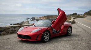 A red sports car with a Tesla badge is parked on a paved coastal road. The driver-side door is open upwards. The ocean