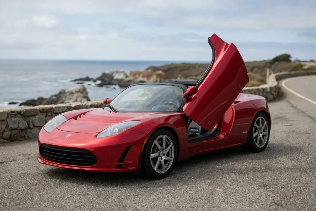 A red sports car with a Tesla badge is parked on a paved coastal road. The driver-side door is open upwards. The ocean